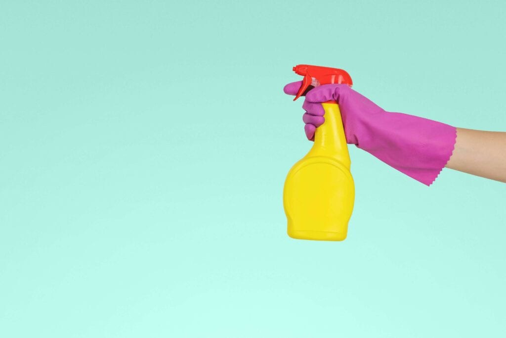 a light blue background and an arm is extending across the screen wearing a pink rubber glove holding a yellow spray cleaner bottle